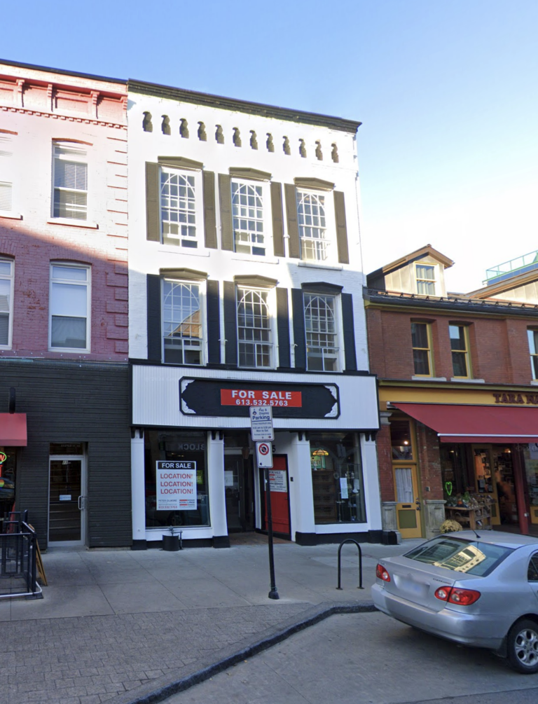 Three storey brick building, with symmetrical fenestration pattern; Sash windows made up of twenty-four panes in the pattern of a Gothic arch; Architectural detailing, including small pediment lintels over each window, the cornice, with corbelled bricks, and brackets, and the small blind arcade; and Central recessed entranceway of double doors and transom, with flanking display windows framed by square wooden columns with brackets at their capitals, supporting the cornice over the first storey.